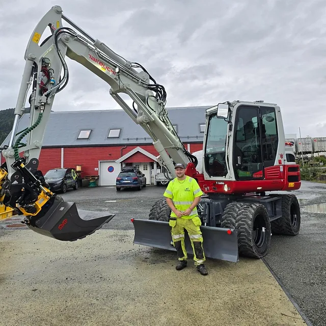 Arbeider foran gravemaskin og rød låve