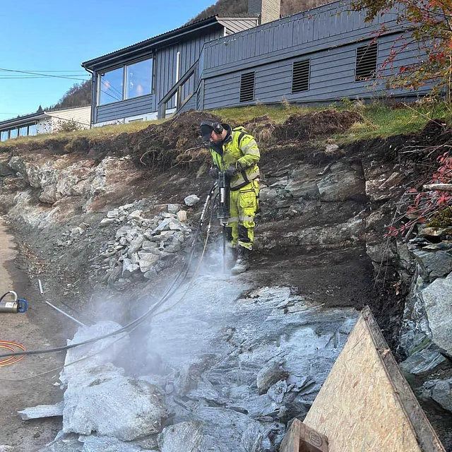 Arbeider i gul vest står ved en fjellskjæring og mur nær et bolighus.