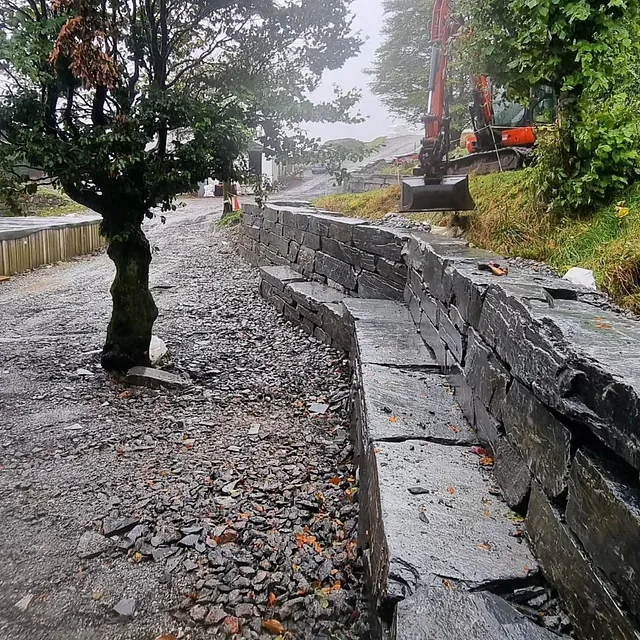 Gravemaskin graver ved kanten av grå steinmur