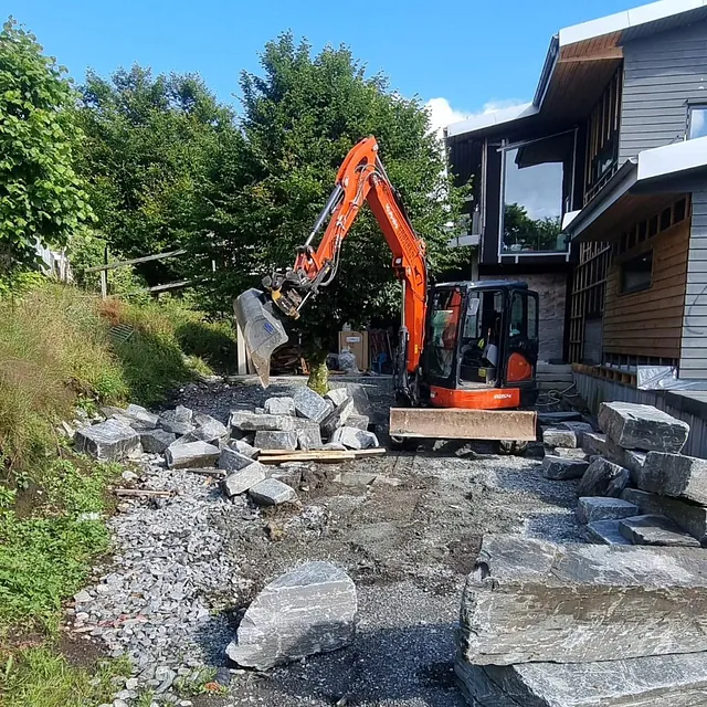 Graving med gravemaskin utenfor grått hus